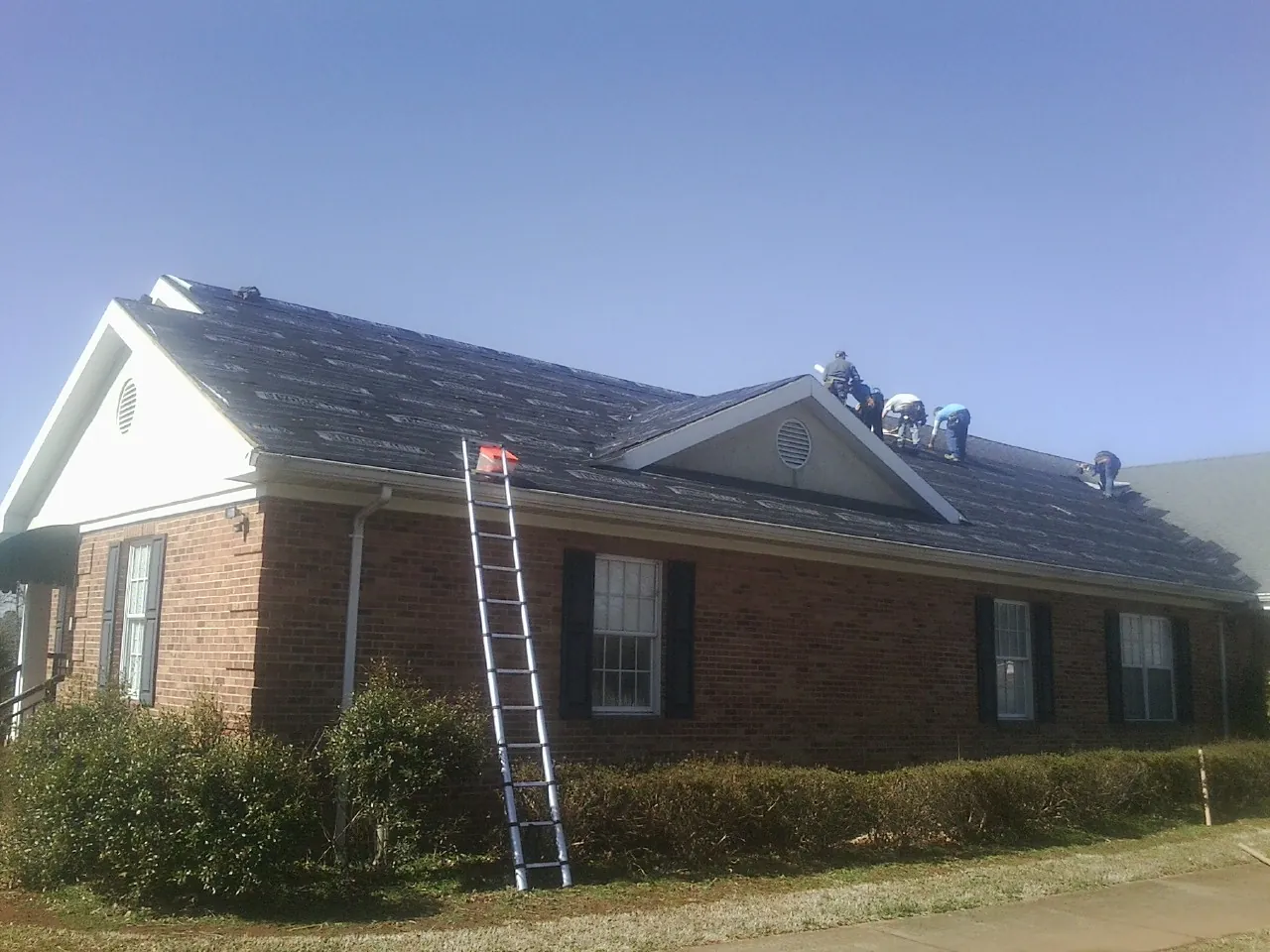 Crew tearing shingles off a brick ranch roof with a ladder set — Emergency Roof Repair work by our crew in La Conner