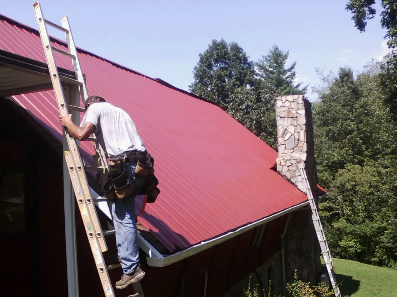 Roofer on a ladder beside a red metal roof and stone chimney — Soffit & Fascia Repair by Roofing Brothers in La Conner